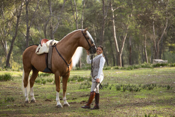 Young woman dressed as a horsewoman, smiling and happy looking at camera next to her beautiful brown horse with white mane, in the middle of a forest. Concept animals, horse riding, equestrian, horse,