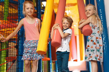 Happy school boy and girls jumping over obstacles in an Indoor playground. Cute kids playing on the colorful playground at a shopping mall. Kids jumping on playground cushions. Children having fun