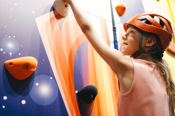 Indoor climbing class for kids. School girl  having fun in indoor playground for children. Happy little girl in red helmet climbing the wall in bouldering center