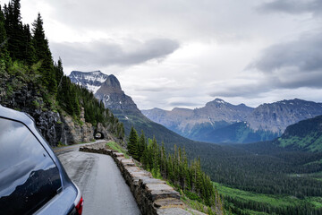 A blue camper style minivan sits at the edge of a natural view of the valley and tall mountain peaks with a road tunnel in the background on Going to the sun road in Glacier National Park Montana