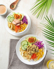 Vegetable salad, Two vibrant bowls filled with pasta, a variety of vegetables, and protein sit on a light tabletop alongside tropical leaves