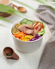 Salad, A vibrant bowl filled with various vegetables, sliced sausage, and fresh greens is displayed on a wooden table, surrounded by natural light
