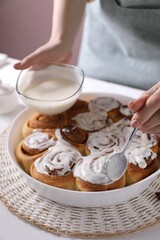 Woman spreading frosting onto freshly baked cinnamon rolls at white table against pink background, closeup