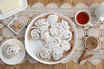 Delicious frosted cinnamon rolls, spices, tea and butter on wooden table, flat lay