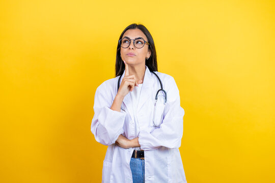 Young brunette doctor woman wearing stethoscope standing over isolated yellow background serious face thinking about question with hand on chin, thoughtful about confusing idea