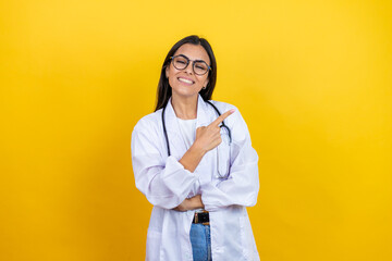 Young brunette doctor woman wearing stethoscope standing over isolated yellow background smiling and pointing with hand and finger to the side