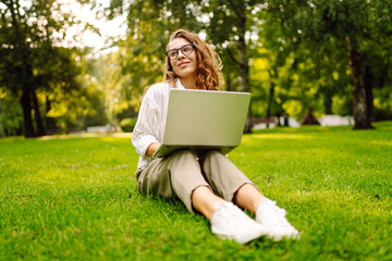 Successful European woman sitting on a green clearing with a laptop. Freelancer, Online, technology concept.