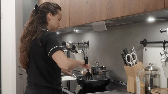 Woman cooks food at home in the kitchen on an induction stove, stirring the ingredients in a pan with a spatula. She fries and cooks the ingredients until fully cooked in a sizzling hot non-stick pan.