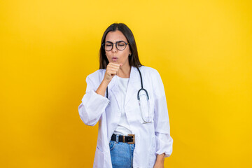Young brunette doctor woman wearing stethoscope standing over isolated yellow background with her hand to her mouth because she's coughing
