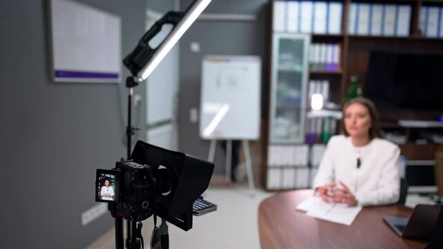 Caucasian brunette wearing white jacket sits at desk talking to camera on tripod. A day-light lamp illuminating the scene. Blog content creation. Blurred backdrop.