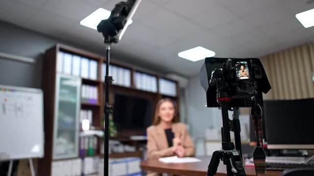 Day-light lamp on the stand and camera on tripod are set in the office. Woman in beige jacket recording blog content. Blurred backdrop.