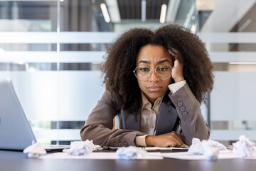 Upset and bored young African American woman in business suit sitting at desk in office, holding head on hand and looking frustrated at crumpled paper