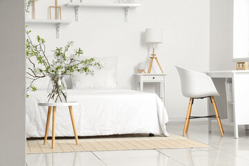 Vase with blooming branches on coffee table near cozy bed in white bedroom