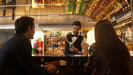 Focusing smiling smart bartender serving gin fizz or tonic soda at luxurious bar, engaging lively conversation with VIP customer couple at back view in atmosphere at lighting warm nightclub. Vinosity.