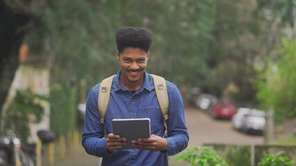 Smiling young man with a backpack holding a digital tablet while walking outdoors. Confident university student enjoying a casual day in nature, ready for remote work or study - Powered by Adobe