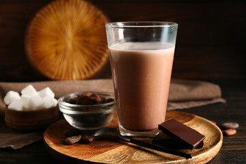 Glass of sweet chocolate milk and bowl with cocoa beans on wooden background