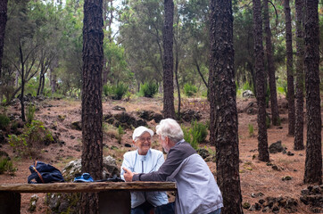 Carefree senior couple sitting at a picnic table in the woods during a hiking day, elderly white haired man and woman enjoying nature