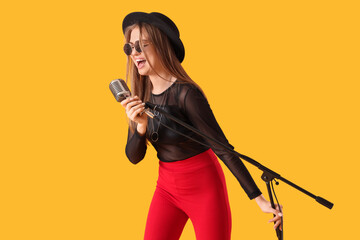 Beautiful young woman with microphone singing karaoke on yellow background