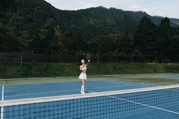Athletic young female tennis player practicing on a vibrant blue court surrounded by lush green...