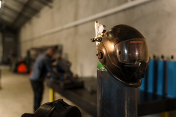 Fototapeta premium Welding helmet hanging on an oxygen or gas cylinder in a workshop with a mechanic working on a vehicle in the background, emphasizing workplace safety and industrial equipment