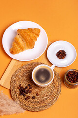 Tasty french croissant on a white plate and a cup of americano coffee from above on an orange background. Sweet breakfast. Morning food.