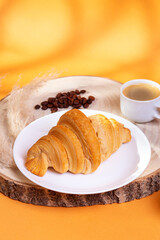 Tasty french croissant on a white plate and a cup of americano coffee in sunlight rays on an orange background. Sweet breakfast. Morning food.