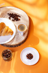 Tasty french croissant on a white plate and a cup of americano coffee in sunlight rays from above on an orange background. Sweet breakfast. Morning food.