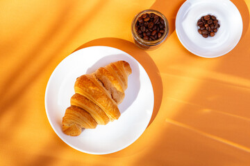 Tasty french croissant on a white plate and a cup of americano coffee in sunlight rays from above on an orange background. Sweet breakfast. Morning food.