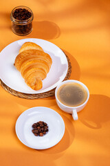 Tasty french croissant on a white plate and a cup of americano coffee in sunlight rays from above on an orange background. Sweet breakfast. Morning food.