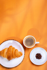 Tasty french croissant on a white plate and a cup of americano coffee in sunlight rays from above on an orange background. Sweet breakfast. Morning food.