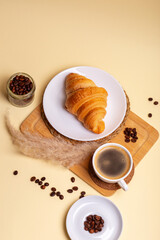 Tasty gold colored french croissant in a white plate, cup of aromatic americano coffee, roasted coffee beans on a wooden desk from above on beige background in rustic style. Sweet breakfast. Morning.