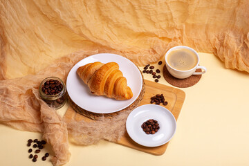 Tasty gold colored french croissant in a white plate, cup of aromatic americano coffee, roasted coffee beans on a wooden desk from above on beige background in rustic style. Sweet breakfast. Morning.