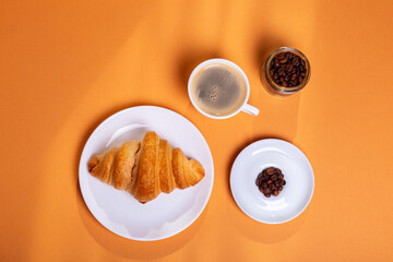 Tasty french croissant on a white plate and a cup of americano coffee in sunlight rays from above on an orange background. Sweet breakfast. Morning food.