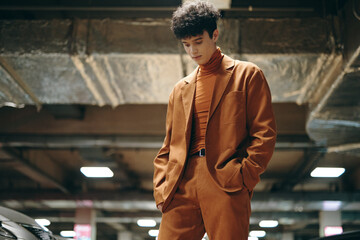 Stylish young man in a brown suit standing in a parking garage, looking down thoughtfully, against a modern urban backdrop