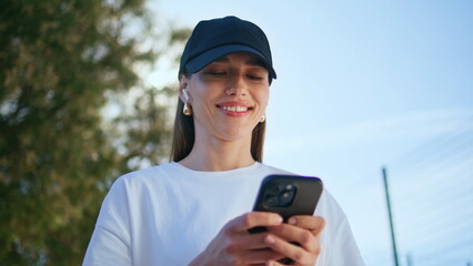 Smiling girl texting smartphone at city street wearing cap closeup. Happy woman © stockbusters