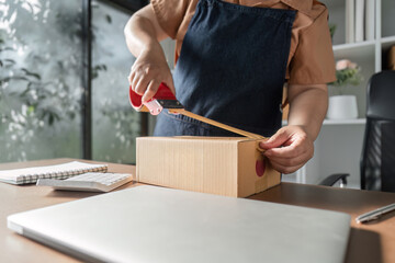 Woman in denim apron taping a cardboard box in a bright home office, preparing it for dispatch