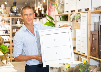 Young man buyer standing in furniture shopping, holding wooden chest of drawers