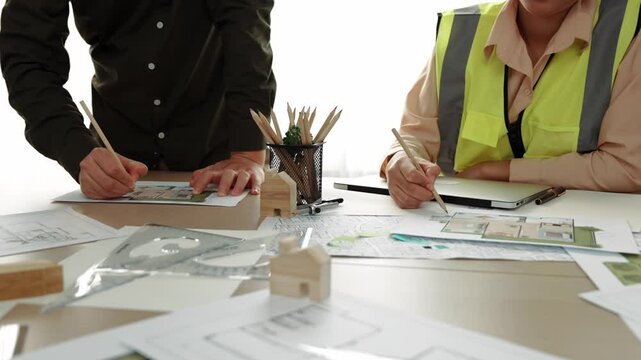 Smart architect and safety engineer discussion about building blueprint on table with architectural document, measurement tools and wooden block scatter around at modern office. Closeup. Delineation.
