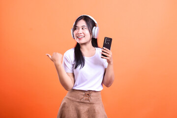 Naklejka premium Young woman with headphones holding phone and pointing to the side on orange background