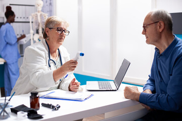 Obraz premium Senior doctor uses digital thermometer on old man to monitor his health in medical consultation. Male pensioner patient waiting at desk for retired female physician to check his temperature in clinic.