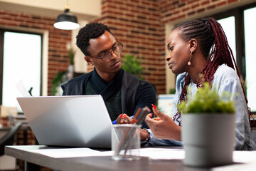 Businesswoman sharing strategy ideas with male employee, having laptop on desk for online reference. African american entrepreneurs sitting with digital device and having discussion in modern office.