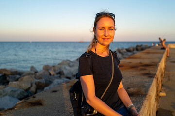 Fototapeta premium Woman sits on a stone wall by the ocean, enjoying the serene sunset view. The calm water and peaceful atmosphere create a beautiful scene.