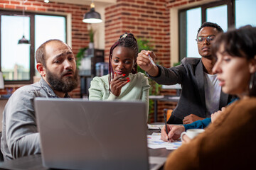 Small group of employees collaborating around a laptop, displaying teamwork in modern office. African american and caucasian colleagues look at device screen, focused on startup project discussions.