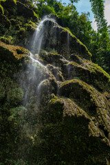 Fototapeta premium Serene waterfall cascading down a moss-covered cliff face, surrounded by lush green foliage.