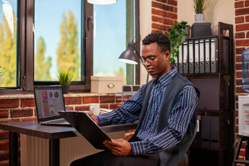 African American professional, sitting at desk near window, reviewing financial paperwork and marketing strategy ideas. Manager with clipboard reading project development plans in brick wall office.