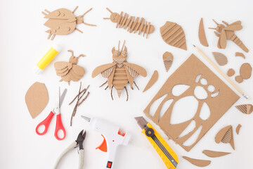 Handmade cardboard insect crafts with crafting tools on white background. Flat lay composition with glue stick, scissors, hot glue gun, utility knife, and cardboard cutouts. Recycling craft,