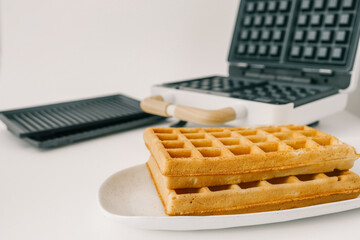 Modern waffle maker with open lid, revealing non-stick black plates with deep square grids. Stylish white body with wooden handles on a neutral background. Kitchen appliance for homemade waffles
