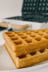 Modern waffle maker with open lid, revealing non-stick black plates with deep square grids. Stylish white body with wooden handles on a neutral background. Kitchen appliance for homemade waffles