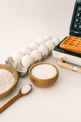 Close-up of waffle-making ingredients with a waffle maker in the background. Wooden bowls with flour and sugar, eggs in a carton, and a golden waffle resting on the machine
