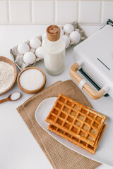 Ingredients for homemade waffles arranged on a white surface, including flour, sugar, eggs, and milk. A waffle maker with wooden handles is visible in the frame